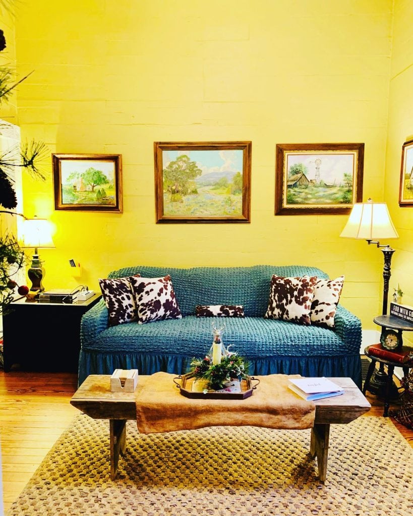 The Cottage at Chappell Hill living room looks straight out of a Hallmark movie—cozy blue sofa, wooden furnishings, antique lighting, and charming scenic décor. Credit: @cottagechtx via Instagram