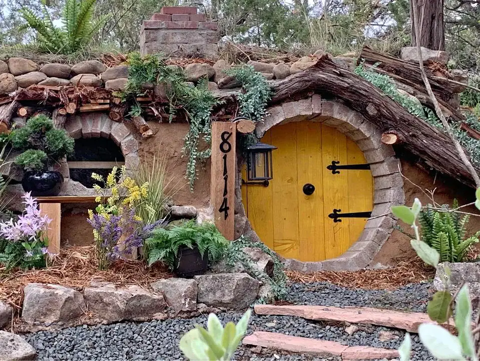 The Hobbit House, with its iconic round wooden door, feels like a portal to another world, surrounded by beautiful rocks, foliage, and blooming flowers. Credit: u/majorvex via r/texas