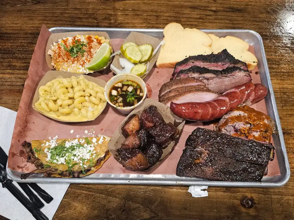 A full spread from Hurtado Barbecue: brisket, sausages, tacos, sides, and those iconic bacon burnt ends — a table that perfectly captures the bold, indulgent spirit of Texas barbecue. Credit: Myra Hennessy