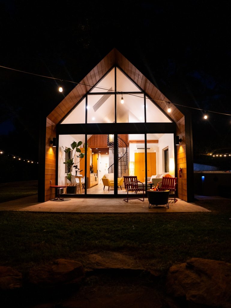 Shadow Bend Cabin glows at night, with floor-to-ceiling glass showcasing the interior, a softly lit patio, and an outdoor seating area ideal for relaxing by the bonfire. Credit: @liveoaklake via Instagram