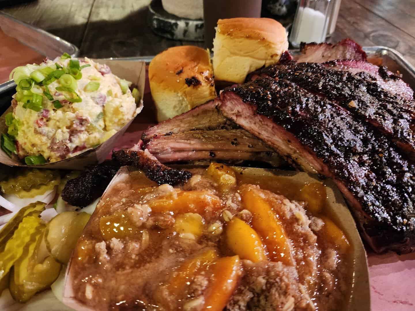 Barbecue spread with beef rib, potato salad, rolls and peach cobbler at Pecan Lodge in Dallas