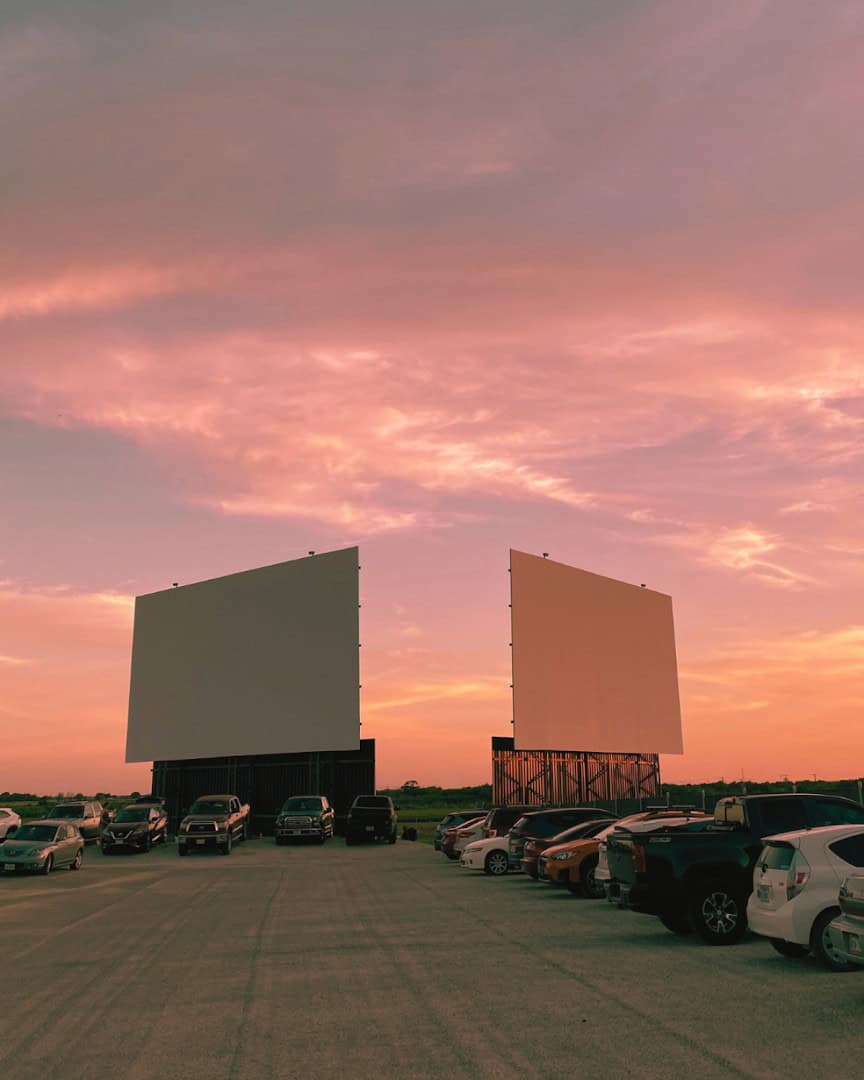 Two drive-in movie screens against a pink sunset with cars parked in New Braunfels, Texas