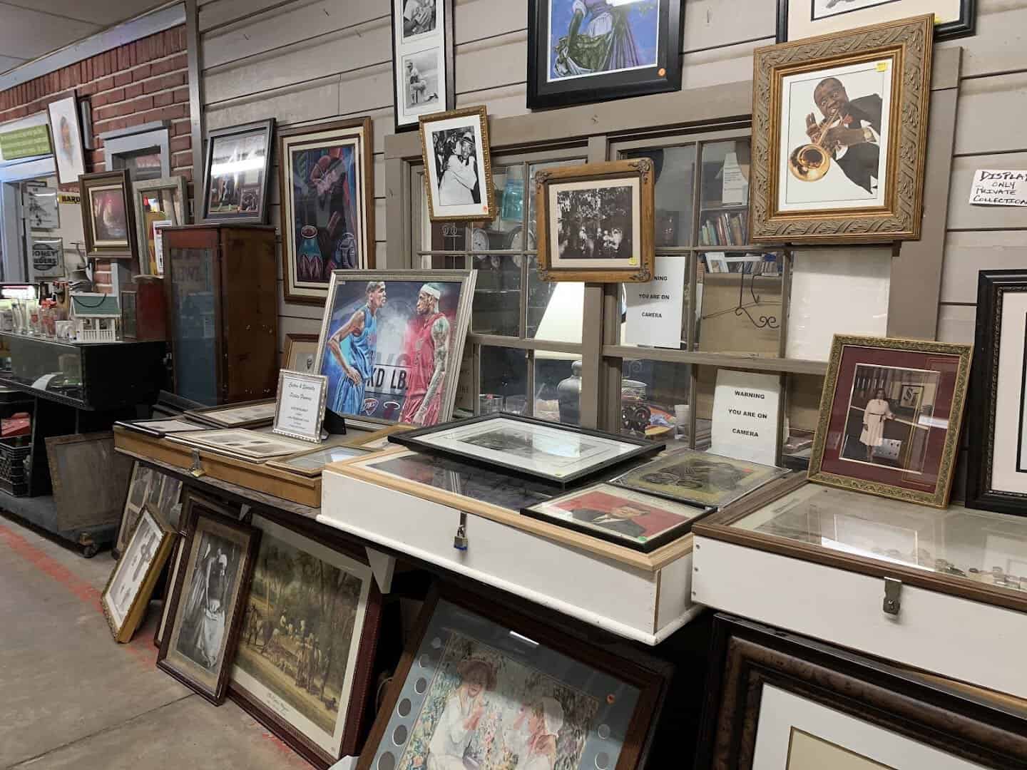 An indoor antiques booth stacked with framed art and a glass display case at Cole's Antique Village in Pearland, Texas