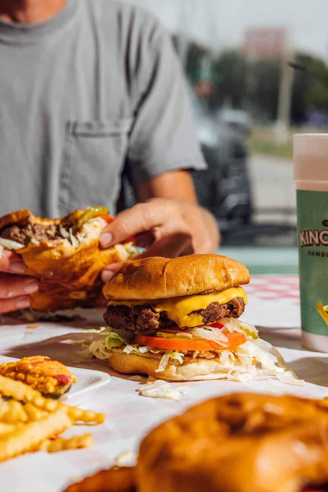 A hand holding a loaded cheeseburger with a side of fries at Kincaid's Hamburgers in Fort Worth