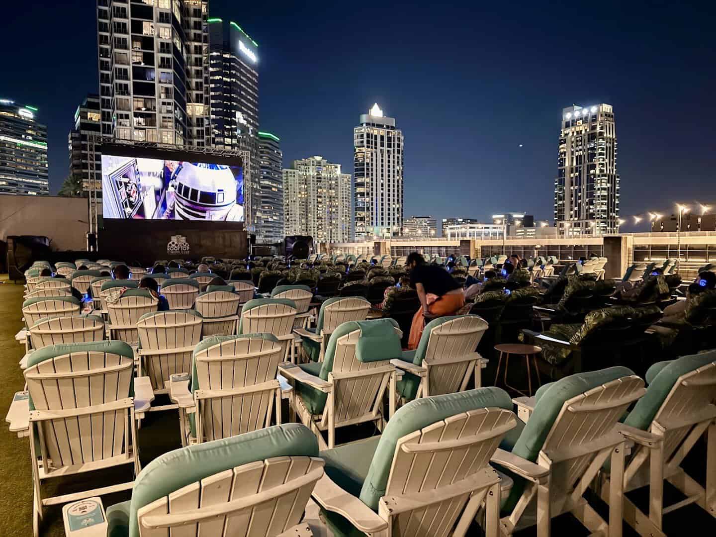 Adirondack chairs with wireless headphones facing the screen and Houston skyline at night at Rooftop Cinema Club Uptown