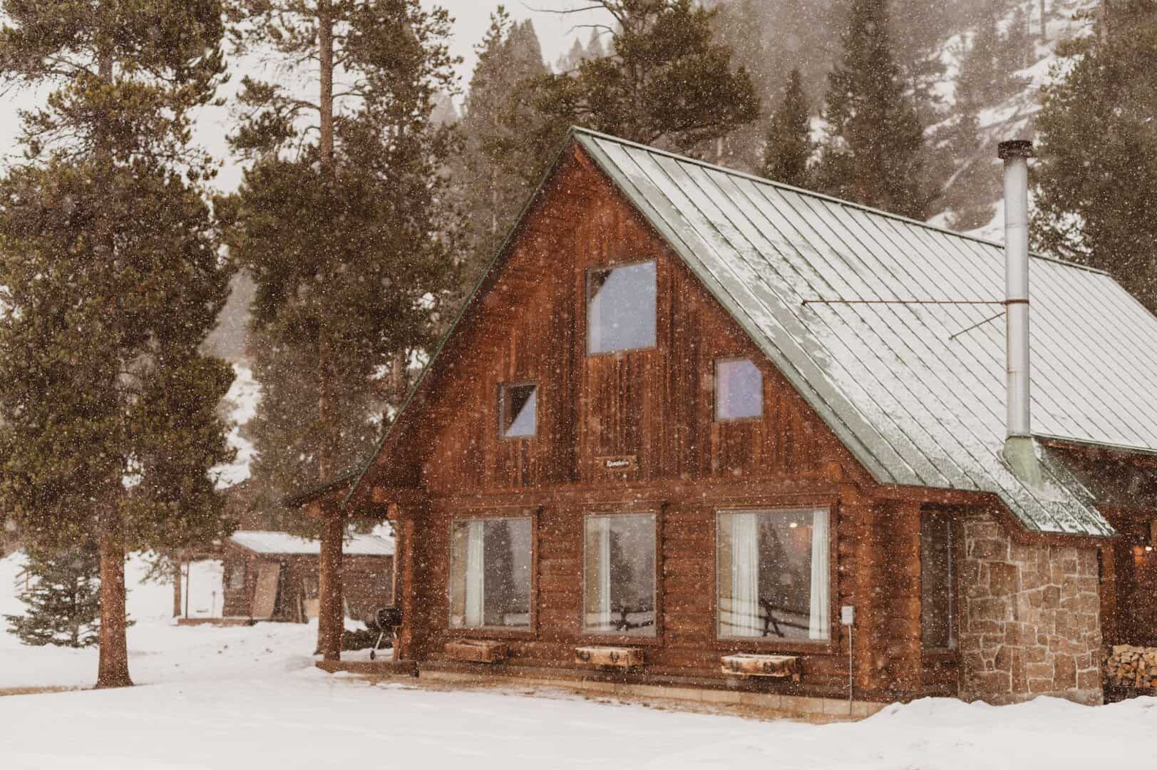 Log cabin with a green metal roof under falling snow and pines, 320 Guest Ranch, Gallatin Gateway, Montana