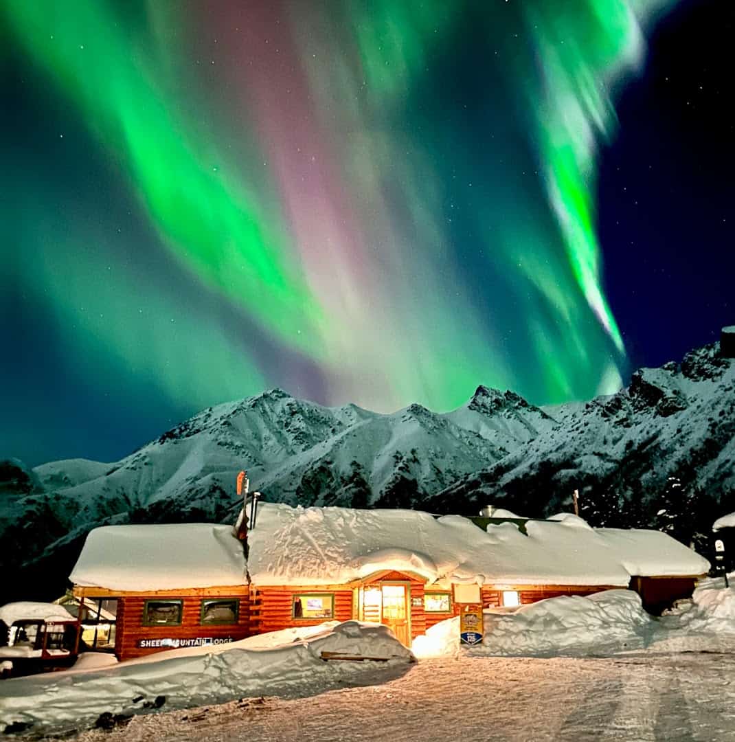 Snow-covered log lodge glowing warm under the northern lights, Sheep Mountain Lodge, Glacier View, Alaska