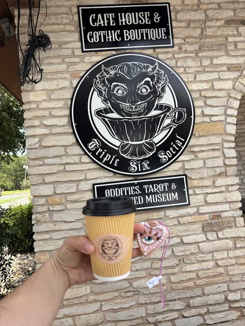 Hand holding a Triple Six Social coffee cup in front of the Cafe House and Gothic Boutique sign in San Marcos, Texas