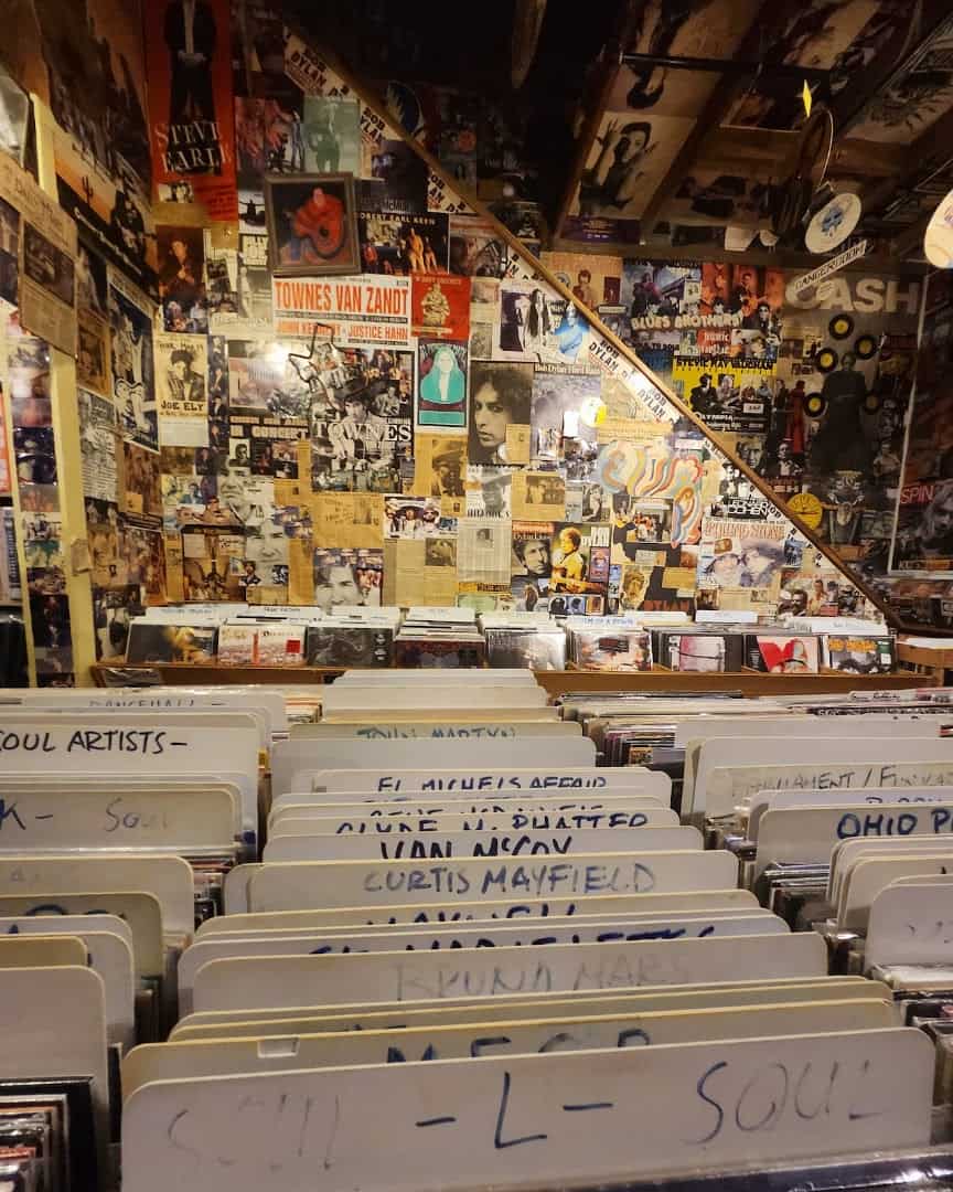 Used record crates with handwritten genre dividers below a wall of clippings at Sigs Lagoon